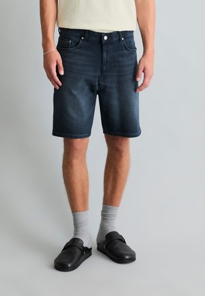 Mężczyzna ubrany w ciemne krótkie spodenki dżinsowe, jasnoszare skarpetki, czarne buty wsuwane i jasną koszulę, stojący na tle jednolitego tła.