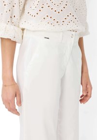 Weiße Hose mit glatter Textur und maßgeschneidertem Design, ausgestattet mit seitlichen Taschen und einem kleinen Metalllogo-Akzent am Bund.