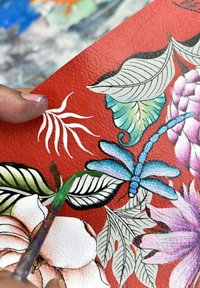 Handbeschilderde leren oppervlak in het rood met ingewikkelde bloem- en bladontwerpen in roze, groen en blauw, met een gedeeltelijk zichtbare kwast.
