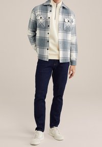 Veste en flanelle à carreaux bleu clair et blanc, dotée de deux poches poitrine et de boutons noirs, portée sur un pull crème et un jean foncé.