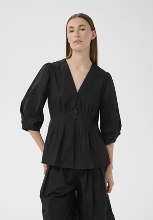 Femme aux longs cheveux raides portant un chemisier noir à col en V boutonné avec des manches bouffantes trois-quarts et un pantalon noir ample assorti.