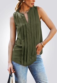 Blusa sin mangas de color verde oliva con pliegues verticales y escote en V. Tejido suave con un corte relajado, combinada con jeans azul claro y un bolso negro.