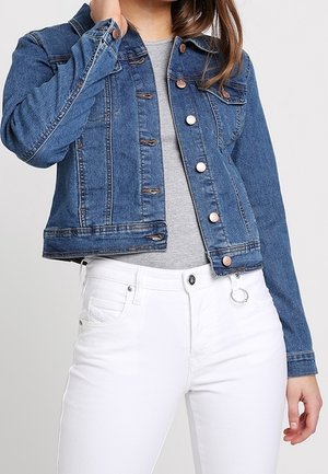 Femme portant une veste en jean bleue par-dessus un top gris et un pantalon blanc, tenant le col de la veste d'une main.