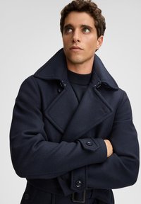 Man met gekruiste armen, gekleed in een donkerblauwe dubbelzijdige jas en een zwarte coltrui, kijkt omhoog en opzij tegen een effen achtergrond.