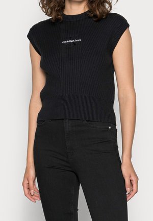 Femme portant un haut noir sans manches en tricot côtelé avec le logo Calvin Klein Jeans et un jean noir taille haute sur un fond clair.