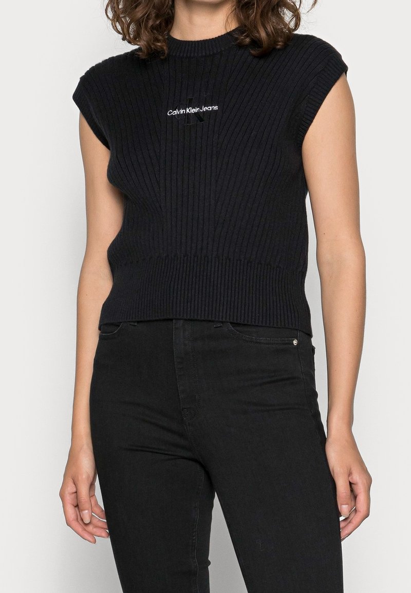 Femme portant un haut noir sans manches en tricot côtelé avec le logo Calvin Klein Jeans et un jean noir taille haute sur un fond clair.
