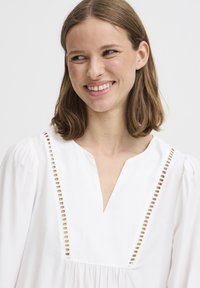 Weiße Bluse mit V-Ausschnitt und offenen Stichdetails entlang des Ausschnitts. Mit lockeren, gepufften Ärmeln und gerafftem Oberteil.