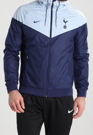 Hombre vistiendo una chaqueta Nike con cremallera de color azul marino y azul claro con el logo de Tottenham Hotspur y pantalones Nike negros.