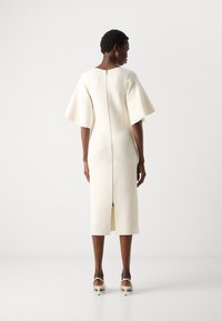 Cream-colored dress with a fitted shape, short bell sleeves, back zipper closure, and a mid-calf length featuring a back slit.