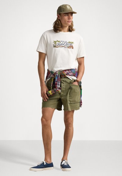 Tricou alb cu mânecă scurtă și grafică cu palmieri, pantaloni scurți verzi măslinie, cămașă colorată cu model legată la талie, încălțăminte bleumarin, capac verde măsliniu.
