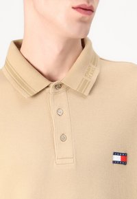 Beige polo skjorte laget av teksturert stoff, med en tre-knappers placket, stripete krage og et lite logo-patch i blått, rødt og hvitt.