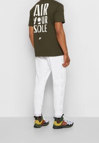 Hombre con camiseta oscura verde "Air Your Sole", pantalones de chándal blancos de Nike con rayas laterales y zapatillas multicolores con cordones y cremalleras.