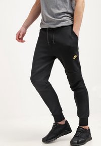 Black Nike joggers with elastic cuffs, featuring a gold logo on the thigh, made from a smooth fabric with a relaxed fit and drawstring waistband.