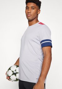 Grey athletic shirt with red accents and blue striped cuffs. Holds a soccer ball with a black and white star pattern.