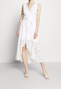 Kvinna i ärmlös vit spetsklänning med hög-låg design, V-ringning och bredt bälte, i kombination med beige vadderade sandaler med öppen tå.