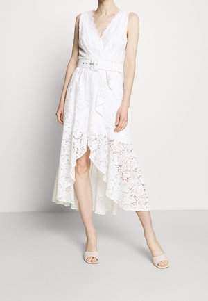 Donna che indossa un vestito bianco in pizzo senza maniche, corto davanti e lungo dietro, con scollatura a V e cintura larga, abbinato a sandali aperti beige trapuntati.