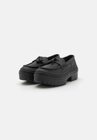 Svarte slip-on sko med tykk plattformsåle, med avrundet design, teksturert overdel og dekorative lisser på en glatt overflate.