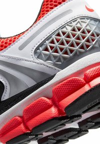 Primo piano di una scarpa da atletica bianca e rossa con tessuto a rete, supporto geometrico in plastica argentata e segmenti di suola in gomma rossa texturizzata.