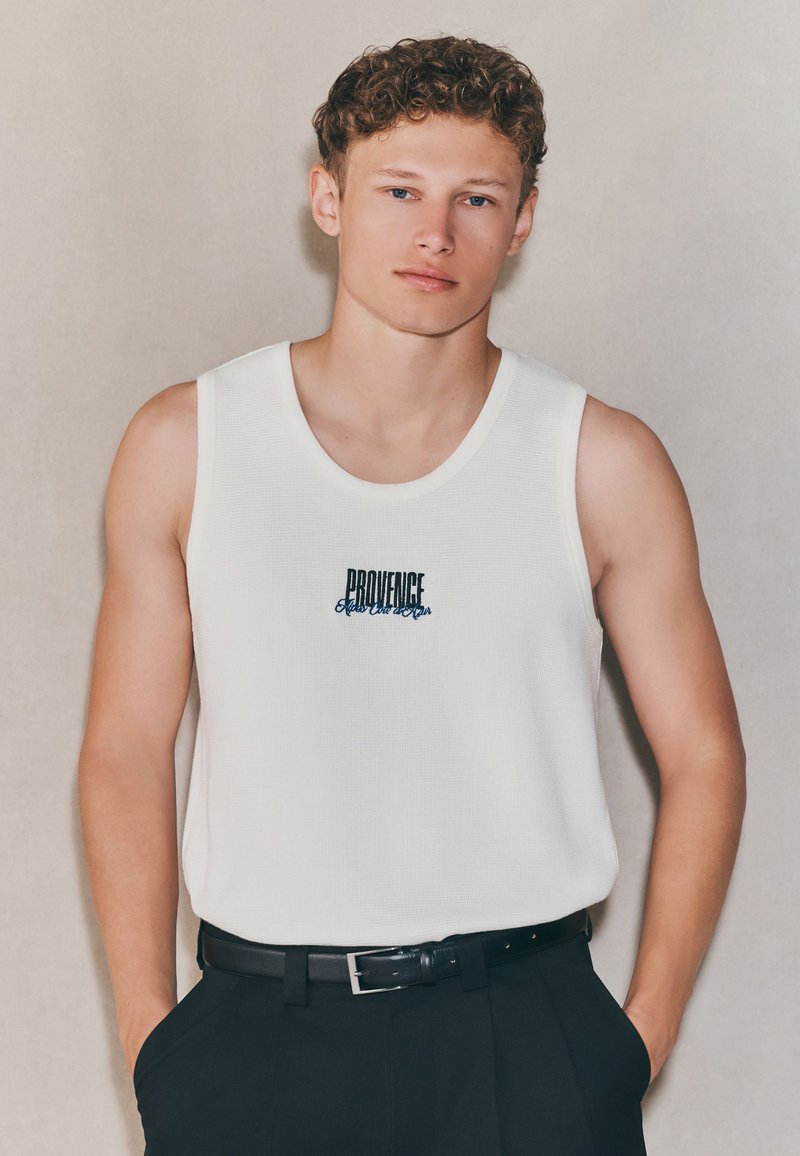 Canotta bianca a coste con la scritta "PROVENCE" ricamata in nero e blu, abbinata a pantaloni neri e una cintura nera con fibbia argento.