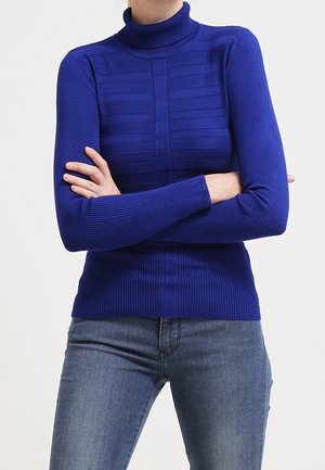 Person wearing a blue turtleneck sweater with horizontal textured stripes and blue jeans, standing with arms crossed against a plain background.