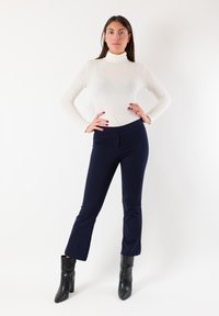Top bianco a collo alto con maniche lunghe abbinato a pantaloni aderenti blu navy e stivali neri con tacco, su sfondo bianco.