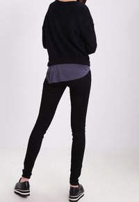 Svart texturerad tröja över en mörkgrå skjorta, tillsammans med svarta skinny jeans och svarta platåskor, sedd bakifrån.