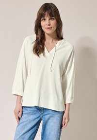 Blouse légère de couleur crème avec un col en V, des cordons de serrage ajustables et des manches trois-quarts. Tissu texturé avec une coupe décontractée.