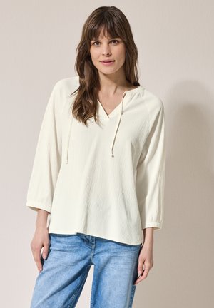 Blouse légère de couleur crème avec un col en V, des cordons de serrage ajustables et des manches trois-quarts. Tissu texturé avec une coupe décontractée.