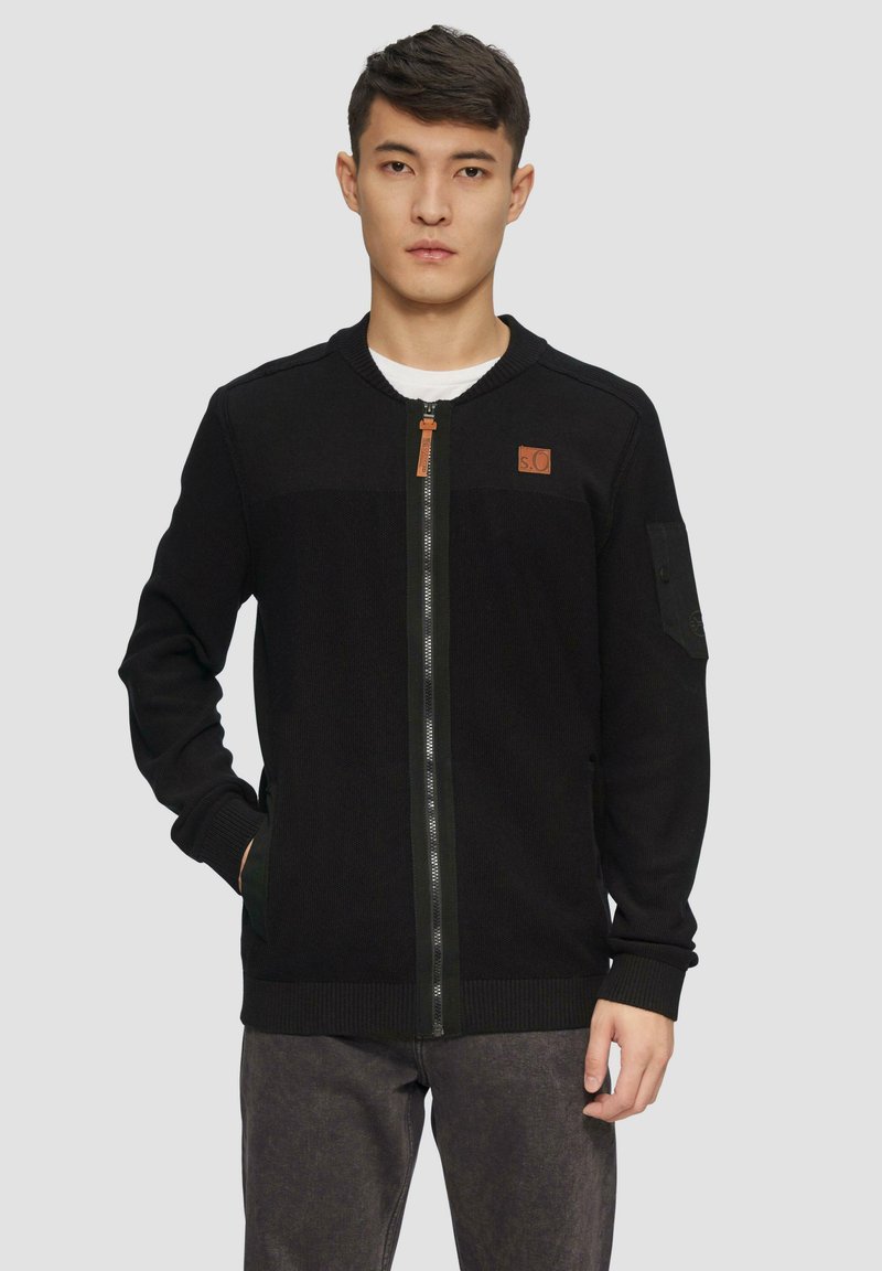 Jonge man in een zwarte ritssluiting cardigan met een klein bruin logo-patch, staand tegen een effen lichtgrijze achtergrond.