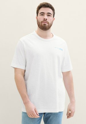 T-shirt blanc à manches courtes en coton, affichant un petit logo bleu sur la poitrine gauche. Coupe ample avec un col rond.