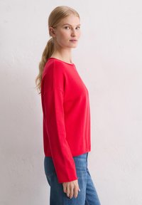 Rotes Langarmshirt mit lässiger Passform, rundem Ausschnitt und strukturiertem Stoff. Kombiniert mit blauen Jeans. Glatte Oberfläche, lebendige Farbe.
