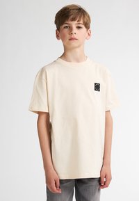 Lichtbeige katoenen T-shirt met korte mouwen en een ronde hals. Voorzien van een klein zwart rond logo op de linkerborst.