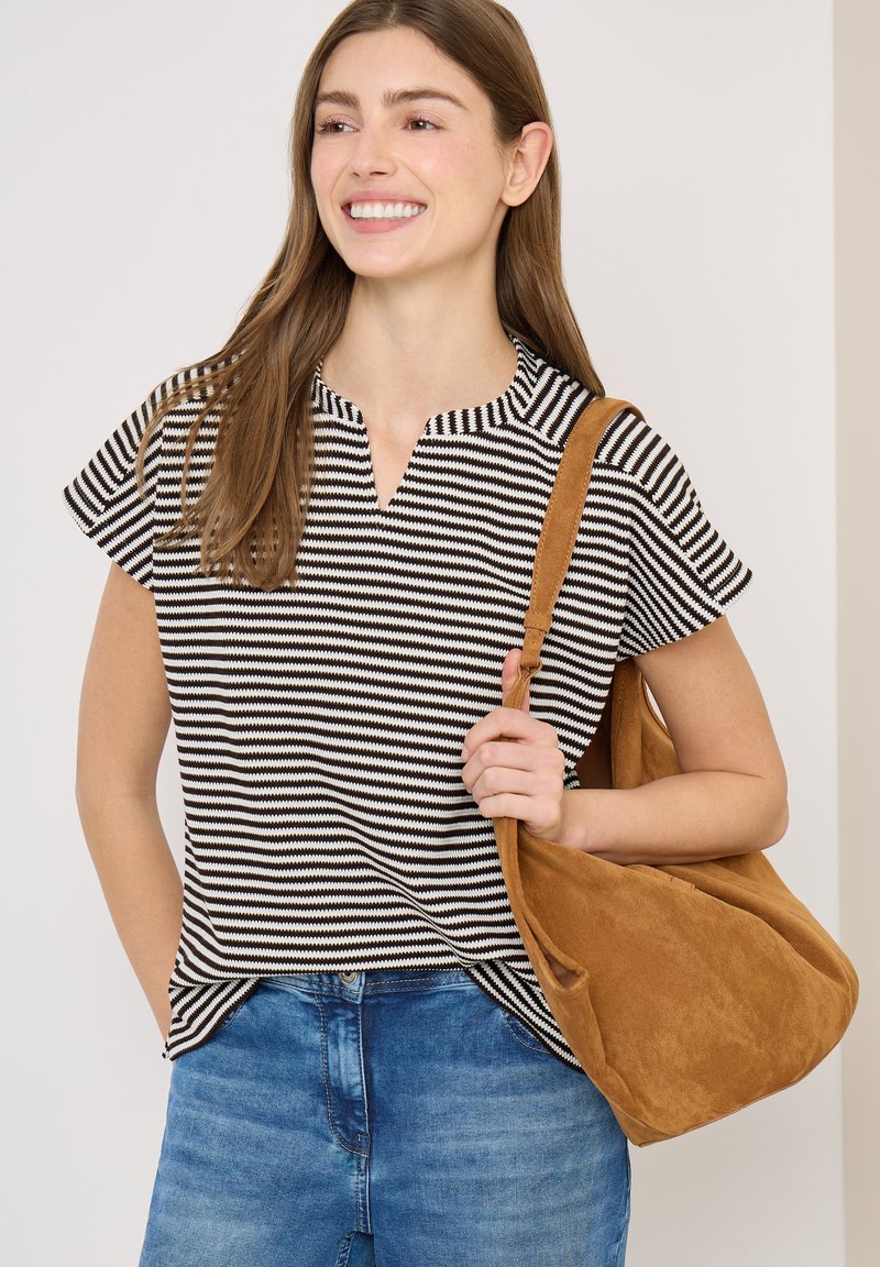 Jeune femme souriante, portant un haut à manches courtes rayé noir et blanc et un jean bleu, tenant un grand sac à bandoulière en daim marron.