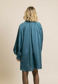 Robe satin teal à manches longues avec un détail froncé au dos. Texture lisse, coupe au genou et poignets resserrés pour plus de silhouette.