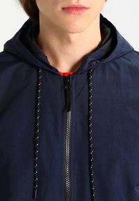 Námořnická kapucínová bunda s zipovým zapínáním a stahovacími šňůrkami se korálkovými detaily. Lehká tkanina s hladkým povrchem.