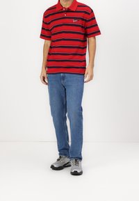 Polo rayé rouge et marine avec un col boutonné et un logo brodé, associé à un jean bleu et des chaussures de sport grises.