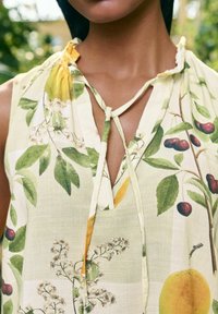 Květinová bezrukávová blůza z jasně žluté látky, s zelenými listy, třešněmi a citrony. Uvázání na krku s volným střihem.