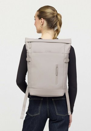 Vrouw met blond gevlochten paardenstaart die een zwart shirt met lange mouwen en een donkere spijkerbroek draagt, en een licht beige rechthoekige rugzak met roll-top meedraagt.