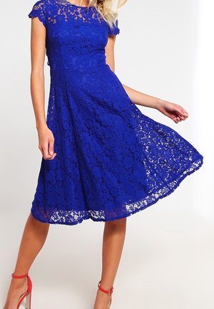 Kvinde iført knælængde kongeblå blondekjole og røde høje hæle med ankelstrop, holder kjolen ud til den ene side.