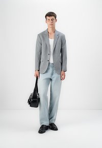 Young man wearing gray blazer, white tank top, light blue jeans, black shoes, holding black leather bag against white background.
