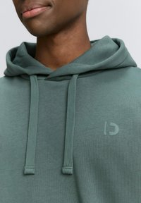 Sudadera verde de tela suave, con un bolsillo en la parte frontal, cordones y un sutil logotipo en relieve en el pecho.