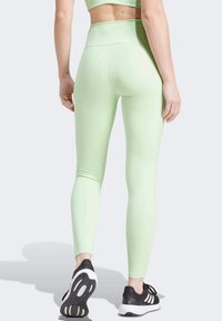 Donna in leggings aderenti color verde chiaro a vita alta e scarpe da ginnastica nere, in piedi con la schiena rivolta verso di noi su uno sfondo chiaro.