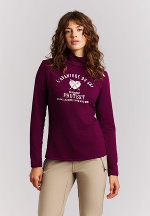 Femme aux cheveux bouclés portant un col roulé violet à manches longues avec un texte blanc sur le thème du ski et un pantalon beige sur un fond uni.