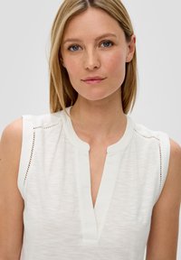 White sleeveless top with a V-neck and subtle textured fabric. Features decorative stitching along the shoulders. Simple and clean design.