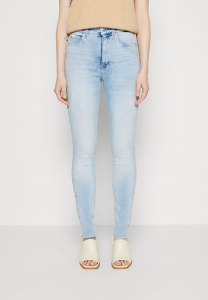 Woman wearing light blue skinny jeans, beige sleeveless top, and cream open-toe mule sandals, standing against a plain white background.