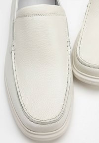 Zapatos de cuero tipo slip-on en gris claro con un acabado texturizado, que presentan costuras en contraste y una suela de goma ligeramente elevada.