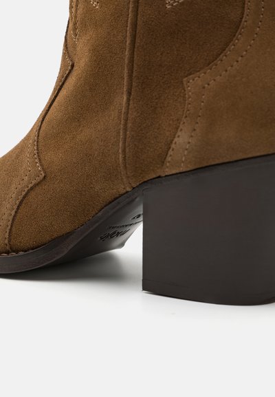 Brūni zamšveida potīšu zābaki ar kvadrāta papēdi, ar izšūšanas akcentiem un gludu tekstūru, parādot sānu profilu.