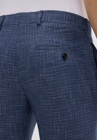 Pantaloni blu con una texture morbida e un motivo intrecciato sottile, dotati di tasche laterali e dettaglio in bottone nero. Tessuto liscio e vestibilità tailored.