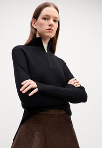 Maglione nero a zip con righe, colletto alto, maniche lunghe e design aderente. Abbinato a una gonna in corduroy marrone.