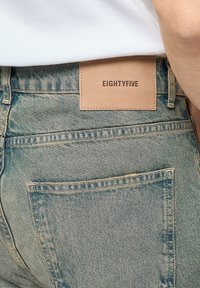 Lyseblå denimjeans med en falmet struktur, forsynet med en beige læderlap mærket "EIGHTYFIVE" på den bageste talje og standardlommer.